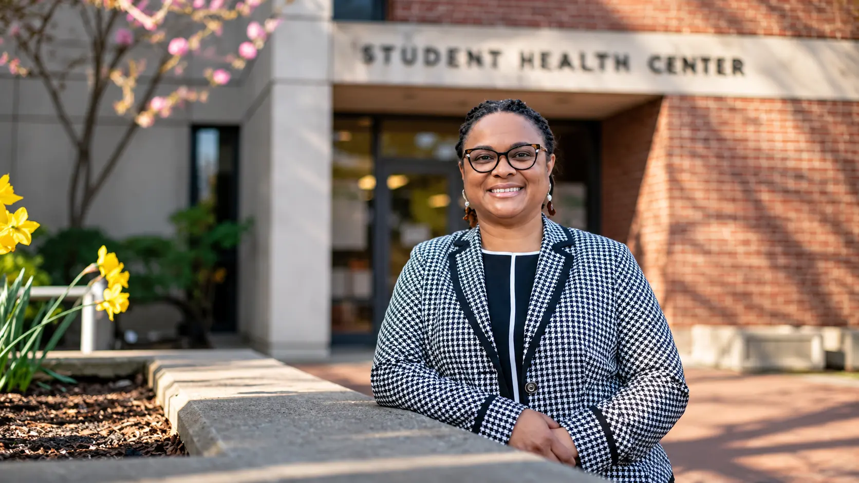 A smiling woman in a houndstooth blazer and glasses stands outside a Student Health Center building.