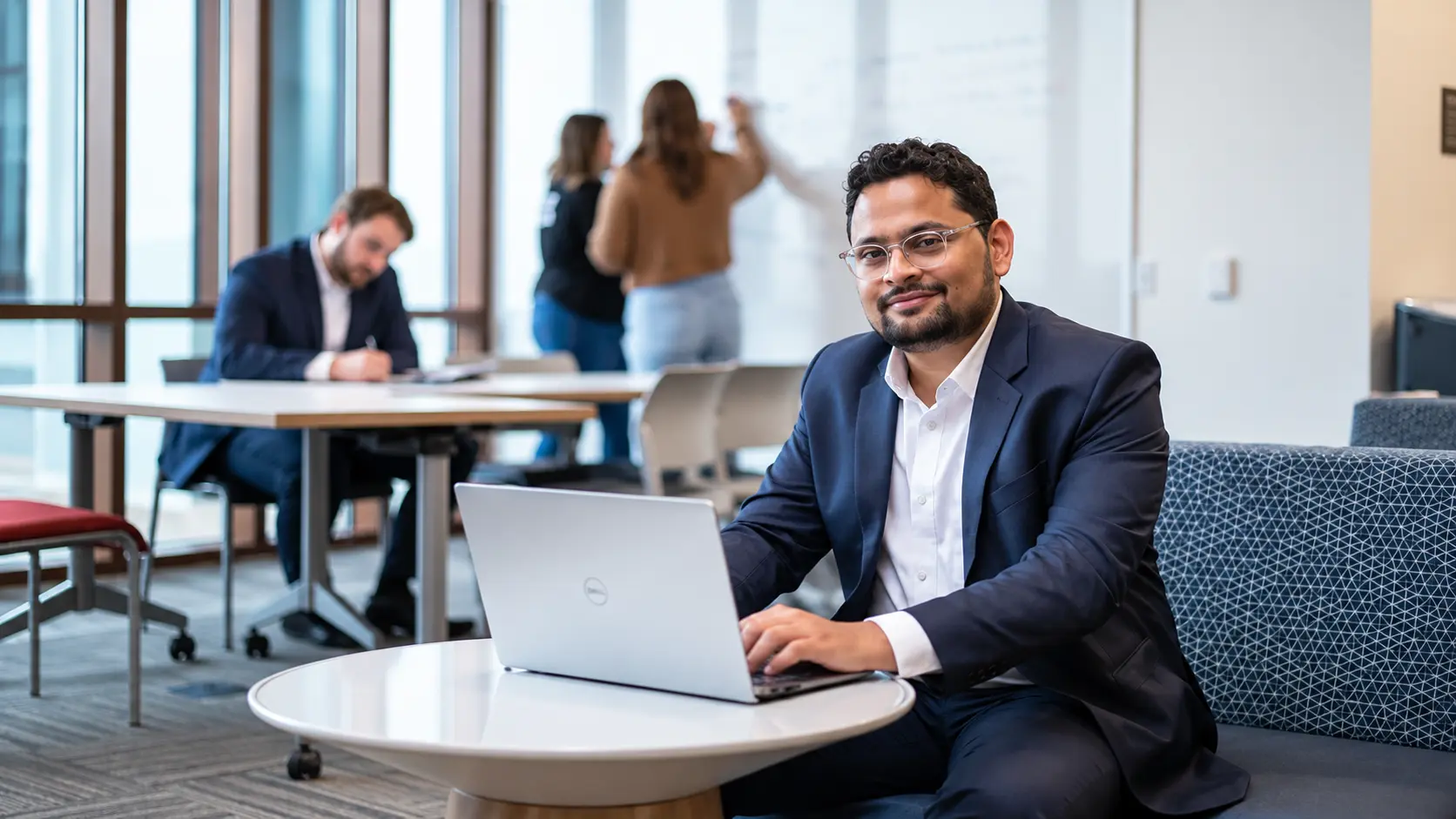 A smiling man in a navy suit works on a Dell laptop in a modern office space.