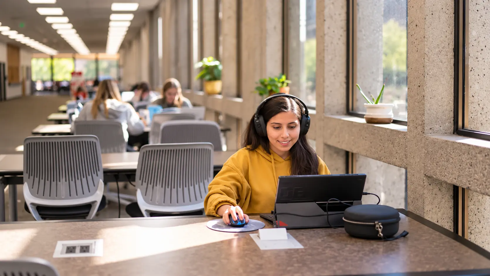 A smiling student wearing headphones and a yellow hoodie uses a laptop in a sunlit library study space.